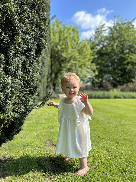 toddler standing in sunny garden wearing white dress in cotton pointelle jersey with frill over shoulders