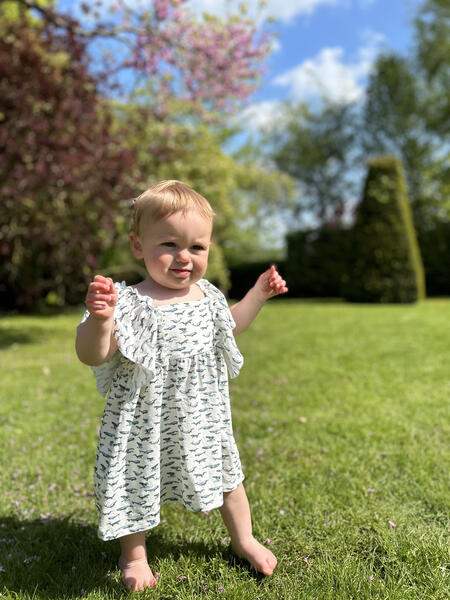 toddler standing in sunny garden wearing white dress in cotton jersey with frill over shoulders in whale print