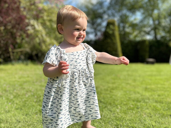 toddler standing in sunny garden wearing white dress in cotton jersey with frill over shoulders in whale print