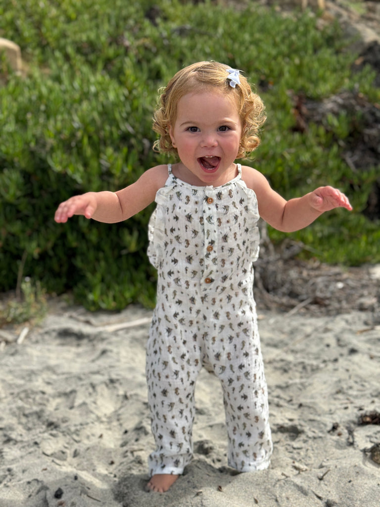 happy toddler on beach in summer sunshine wearing jersey long legged romper with shoe string straps and frills Small seahorse print on white background