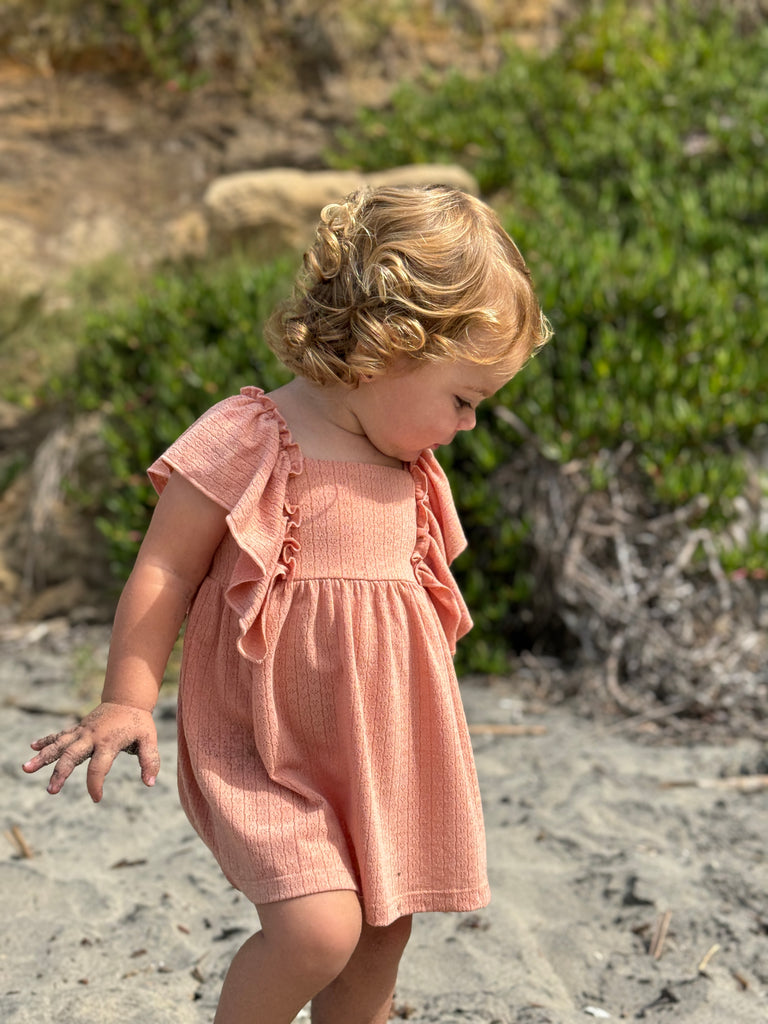 toddler on beach on sunny day wearing coral dress in pointelle jersey with frill over shoulders