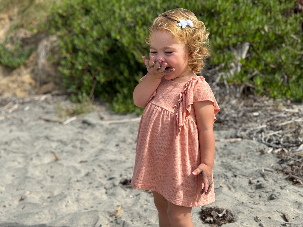 happy toddler laughing on beach on sunny day wearing coral dress in pointelle jersey with frill over shoulders