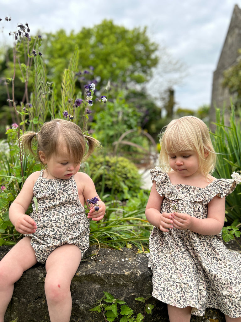 toddler sat on stone wall on sunny day wearing cream bubble with shoe string straps in flower and bird print with friend wearing dress in matching print