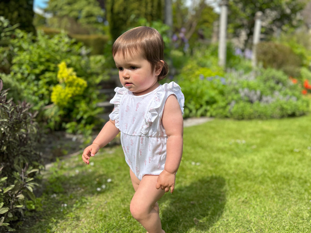 happy baby stood in summer garden wearing jersey cotton romper with frill detail. pink seahorse all over print. short sleeve and legs