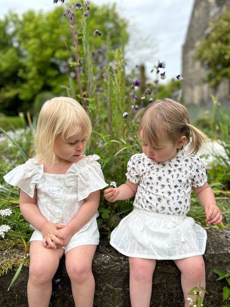 two toddlers sat on stone garden wall one wearing delicate white skort and white seahorse print top and the other matching white shortie romper