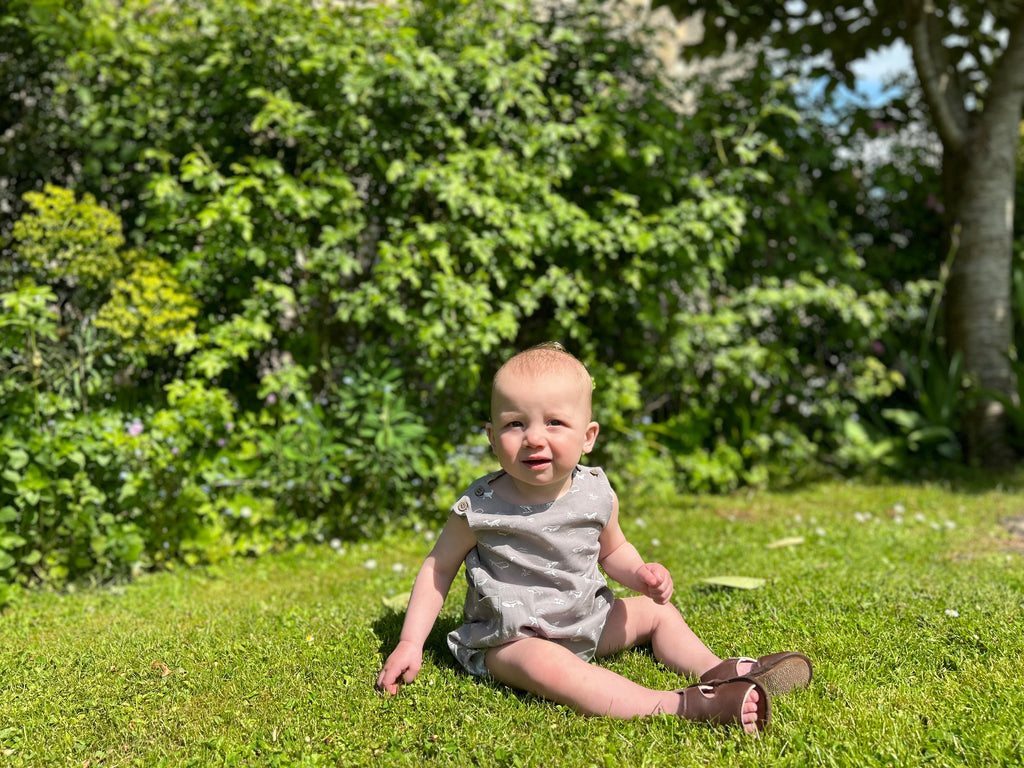 baby boy sat on grass in sunny garden wearing bubble romper in grey with whale print all over 2 buttons on each shoulder strap elasticated leg holes