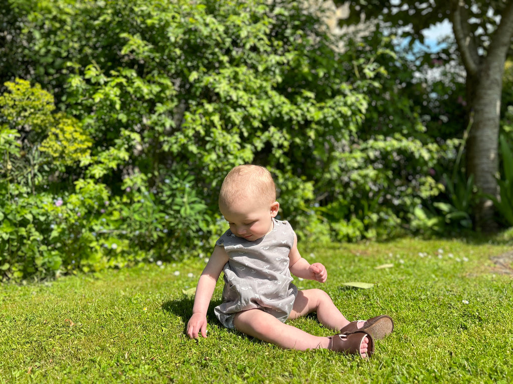 baby boy sat on grass in sunny garden wearing bubble romper in grey with whale print all over 2 buttons on each shoulder strap elasticated leg holes