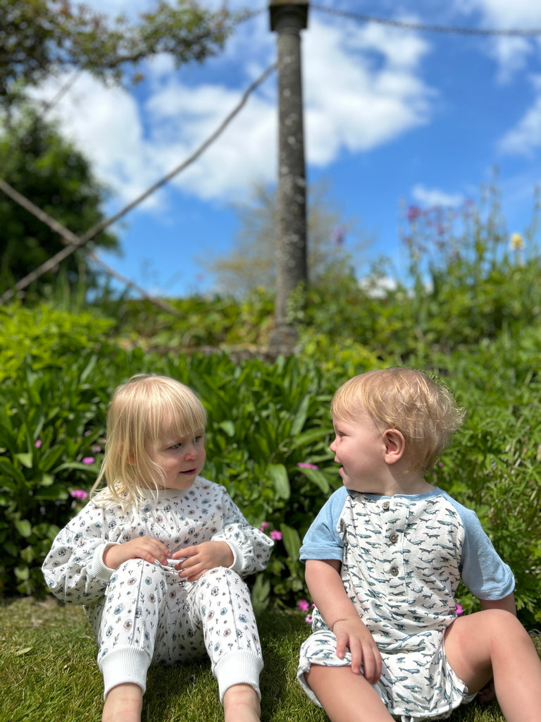 happy toddler sat in sunny garden wearing white sweatshirt with pastel flower print and matching pants sitting next to baby boy in matching romper