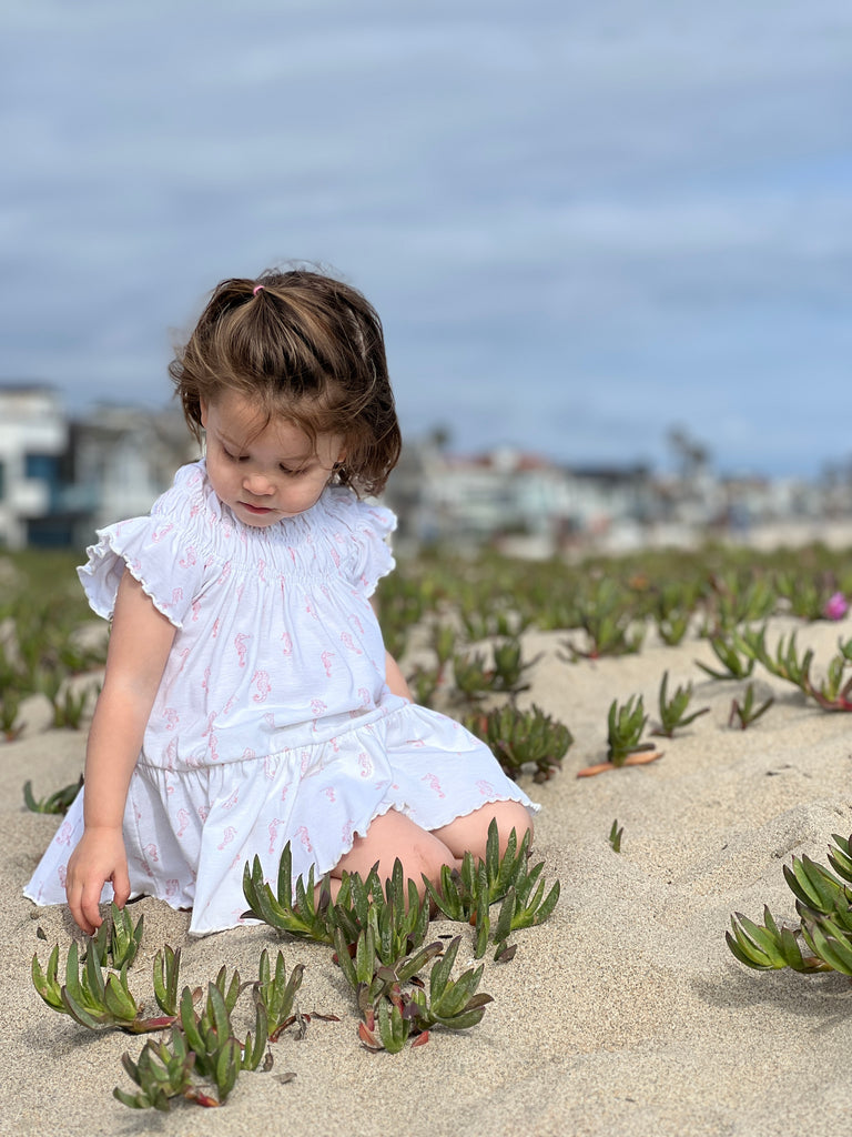 girl sat on beach on summer day wearing white dress with pink flower print smocked neck line short sleeves and large frill tier