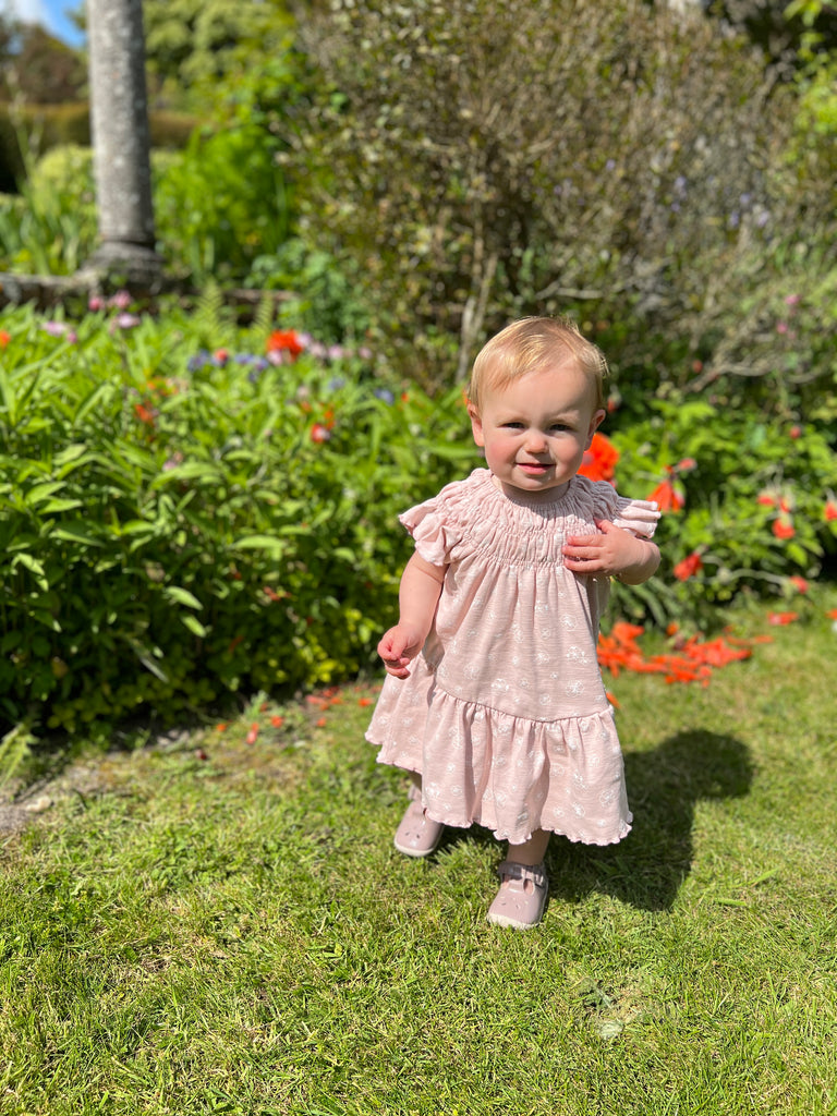 toddler standing on grass in sunshine on summer day wearing pale pink dress with white flower print smocked neck line short sleeves and large frill tier