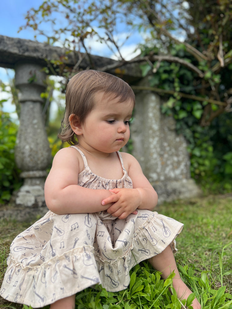 toddler sat in summer garden  wearing cream dress with guitar print elastic waist shoestring shoulder straps hem frill