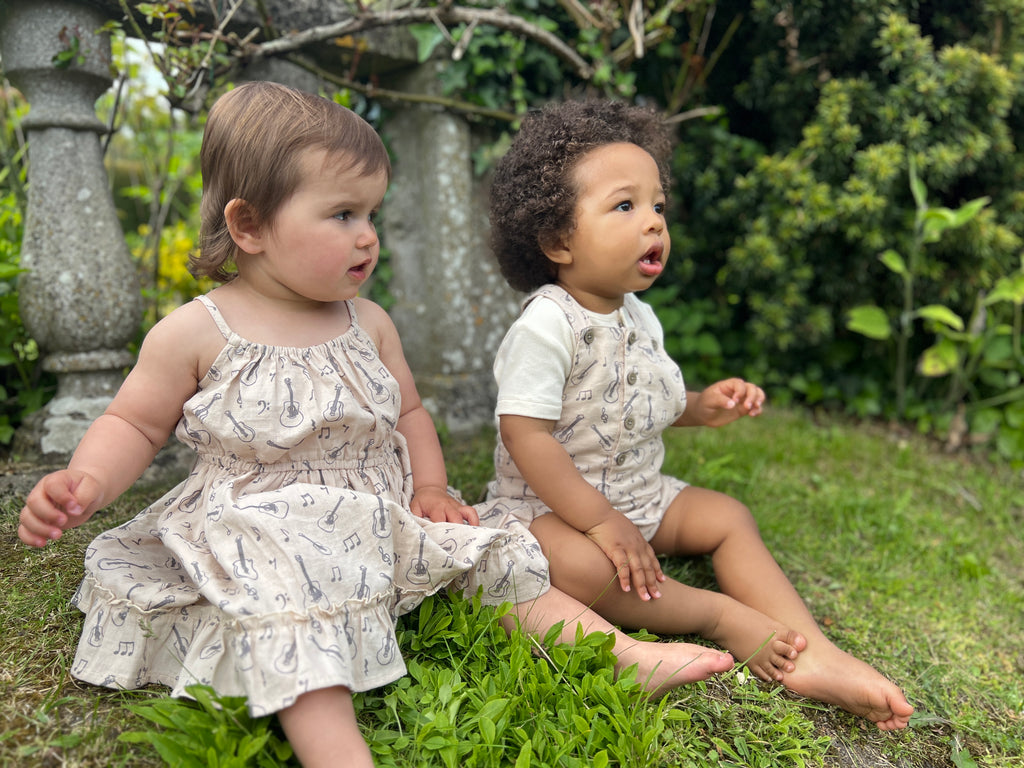 toddler sat in summer garden on sunny day wearing cream dress with guitar print elastic waist shoestring shoulder straps hem frill next to boy in coordinating overalls