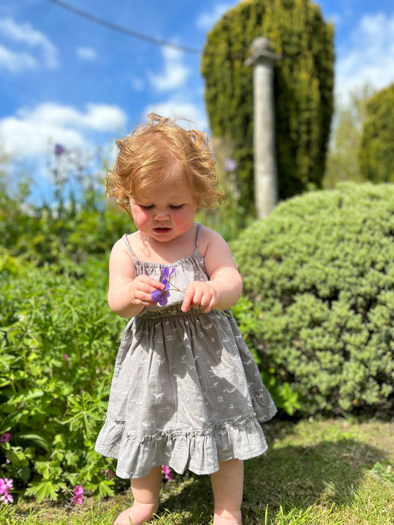 toddler in garden on sunny day wearing grey dress with whale print elastic waist shoestring shoulder straps hem frill