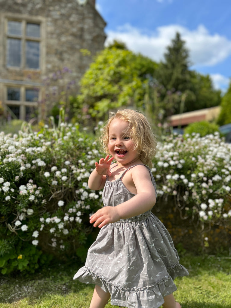 happy toddler in garden on sunny day wearing grey dress with whale print elastic waist shoestring shoulder straps hem frill