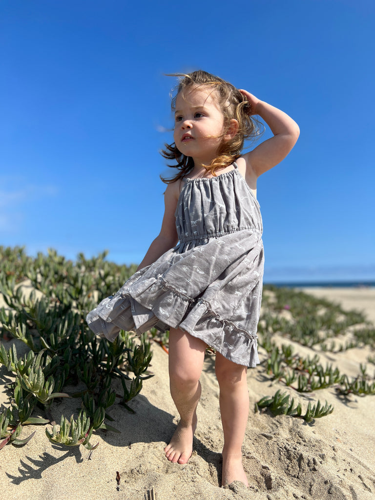 girl stood on beach on sunny day wearing grey dress with whale print elastic waist shoestring shoulder straps hem frill