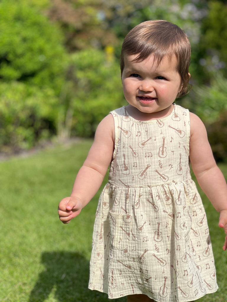 toddler in sunny garden wearing cream gauze sleeveless dress with all over brown guitar print 2 front patch pockets