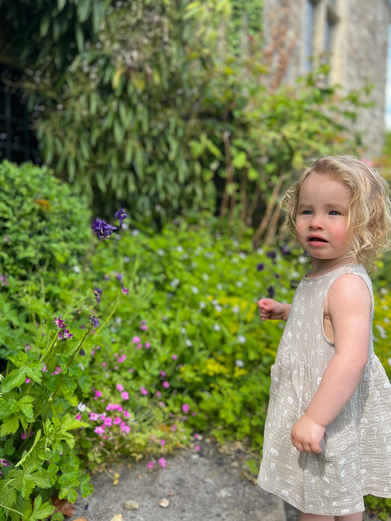 toddler in front of old stone manor house with garden plants wearing grey gauze sleeveless dress with all over white gardening image print 2 front patch pockets