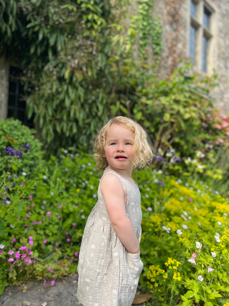 toddler with hands in pockets in front of old stone manor house with garden plants wearing grey gauze sleeveless dress with all over white gardening image print 2 front patch pockets