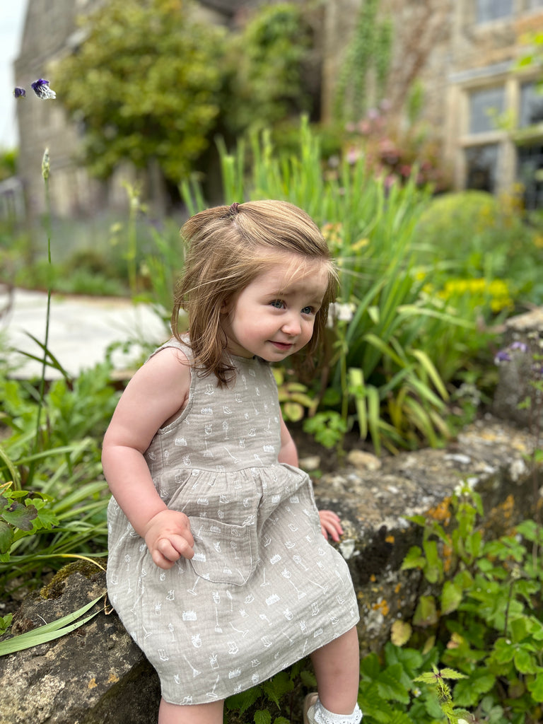 toddler sat in front of old stone manor house with garden plants sitting on old stone wall wearing grey gauze sleeveless dress with all over white gardening image print 2 front patch pockets