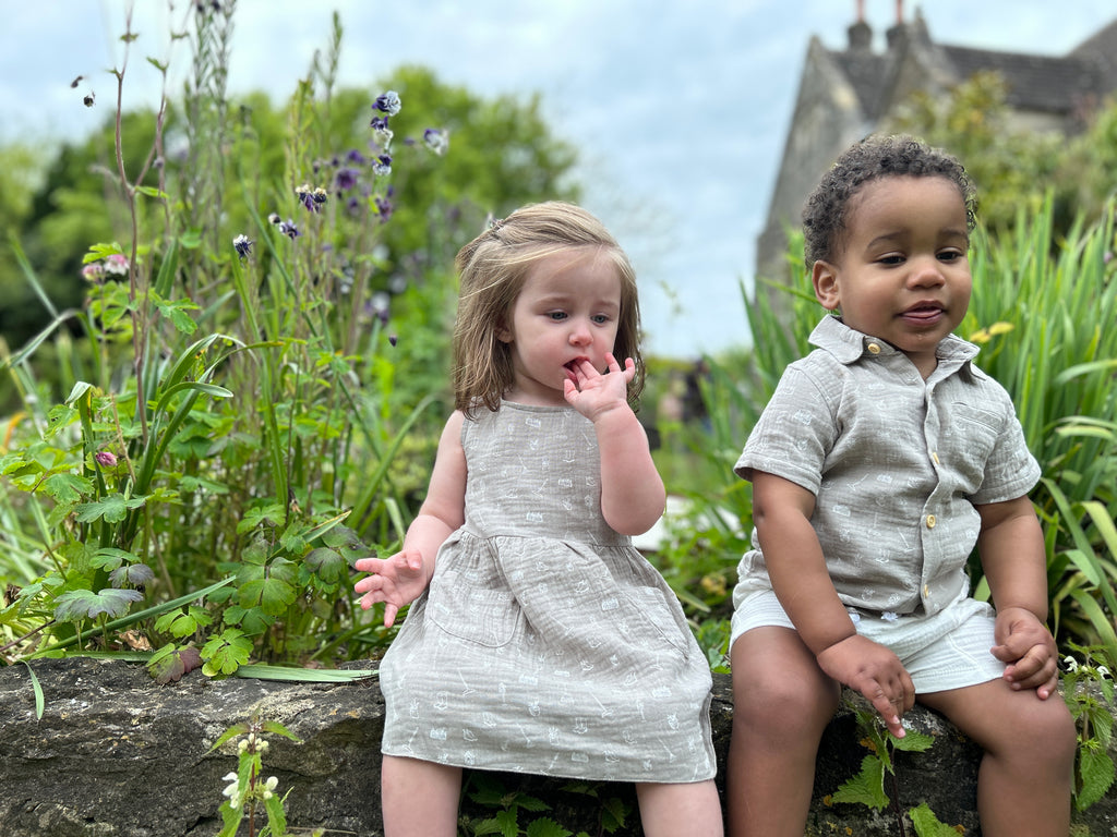 two toddlers sat in front of old stone manor house with garden plants sitting on old stone wall girl wearing grey gauze sleeveless dress with all over white gardening image print 2 front patch pockets boy wearing matching shirt and white gauze shorts