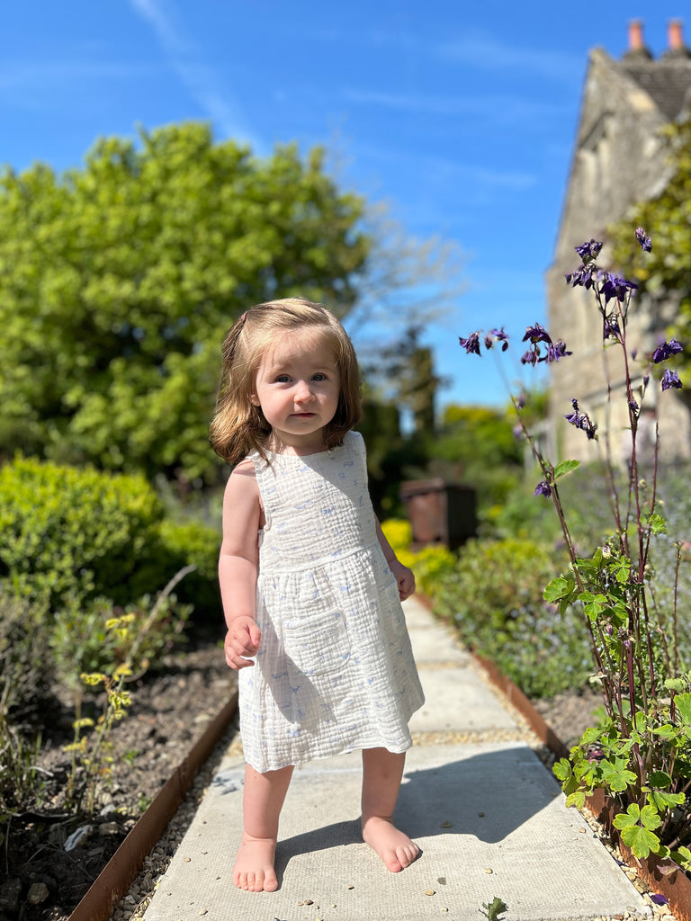 happy toddler standing on garden path in between summer flowers on sunny day wearing white gauze sleeveless dress with all over blue whales print 2 front patch pockets