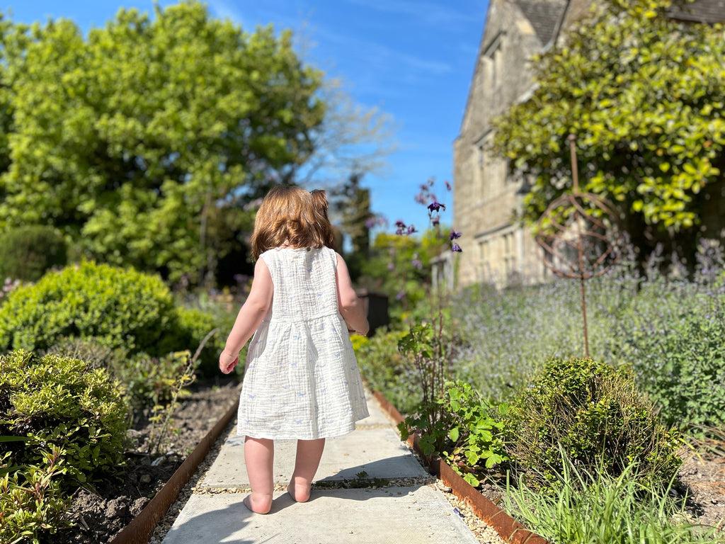 back view of toddler standing on garden path in between summer flowers on sunny day wearing white gauze sleeveless dress with all over blue whales print 2 front patch pockets