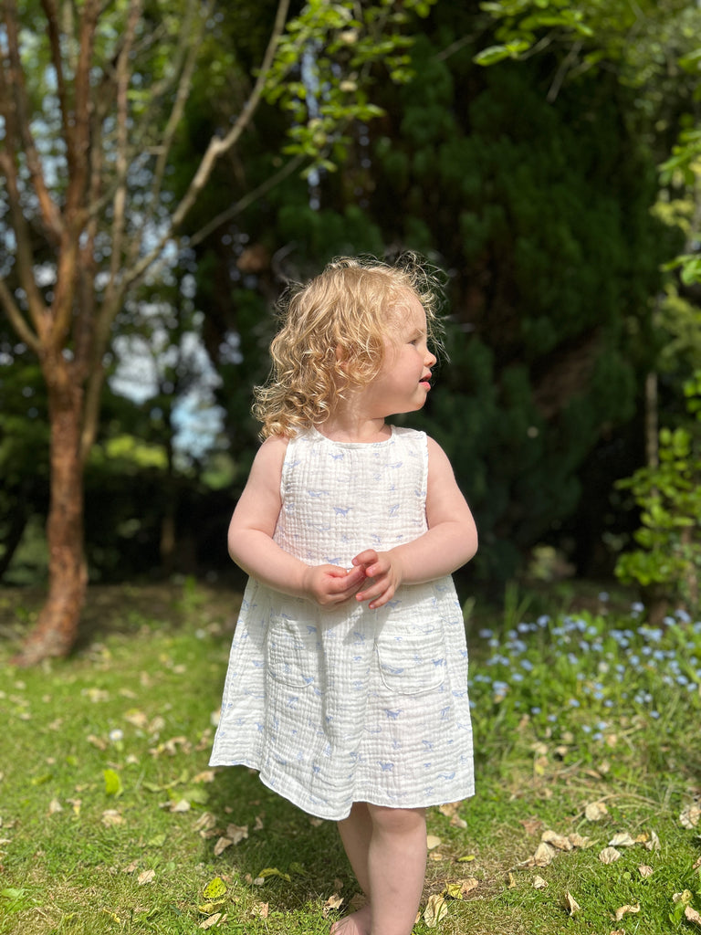 happy toddler standing on grass on sunny day wearing white gauze sleeveless dress with all over blue whales print 2 front patch pockets