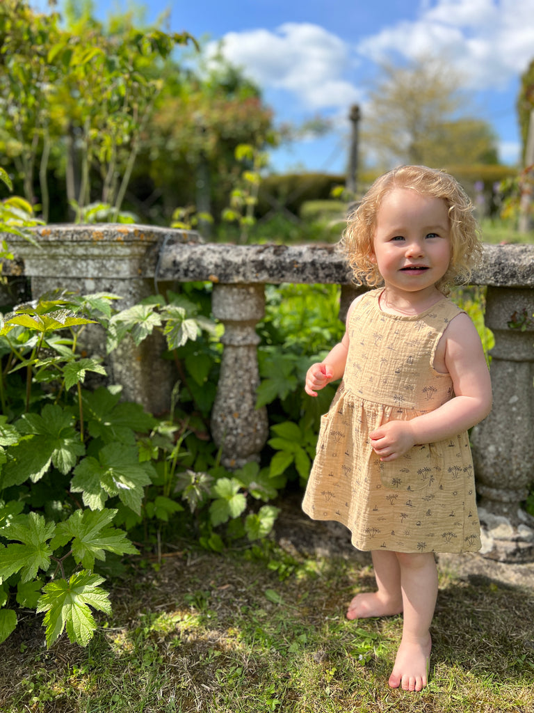 happy toddler stood in front of old stone garden wall wearing cream gauze sleeveless dress with all over island, boat and palm tree print 2 front patch pockets