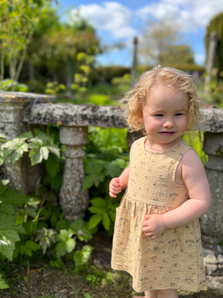 happy toddler stood in front of old stone garden wall wearing  cream gauze sleeveless dress with all over island, boat and palm tree print 2 front patch pockets