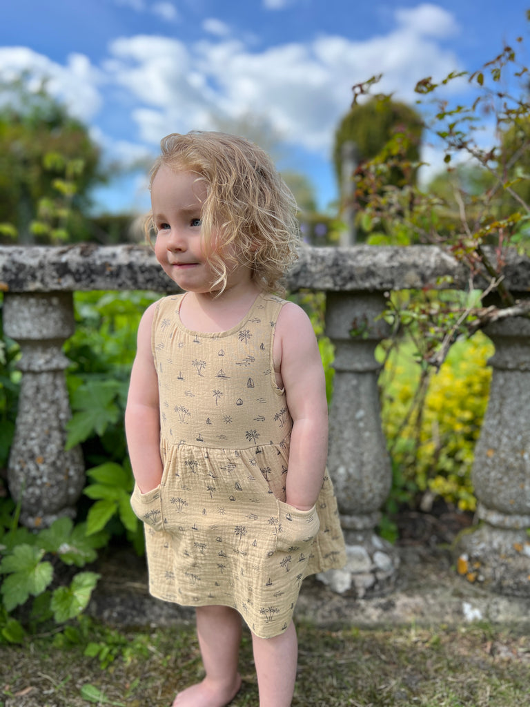 happy toddler with hands in pockets stood in front of old stone garden wall wearing cream gauze sleeveless dress with all over island, boat and palm tree print 2 front patch pockets