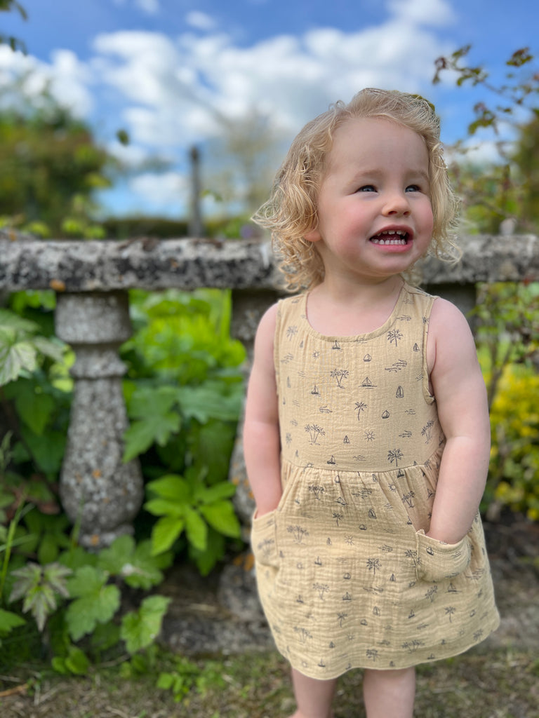 happy toddler with hands in pockets stood in front of old stone garden wall wearing cream gauze sleeveless dress with all over island, boat and palm tree print 2 front patch pockets