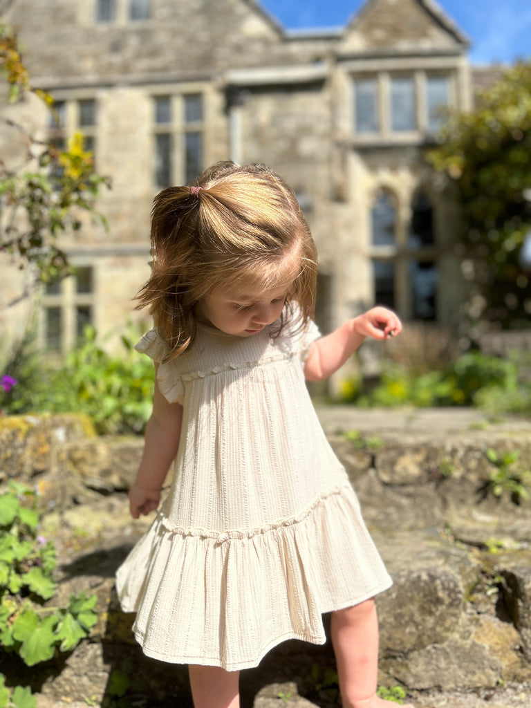 happy toddler standing in front of old stone manor house wearing cream two tiered frill dress with frill sleeves