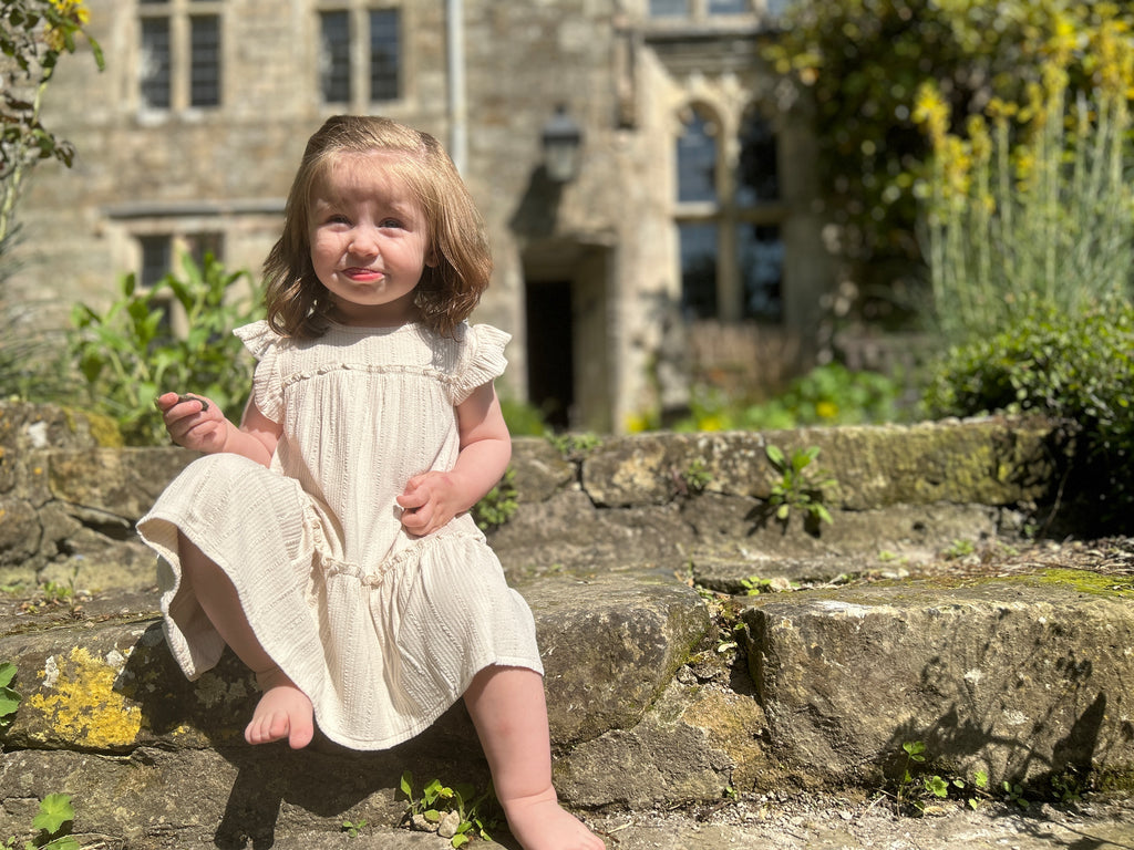 happy toddler sat in front of old stone manor house wearing cream two tiered frill dress with frill sleeves