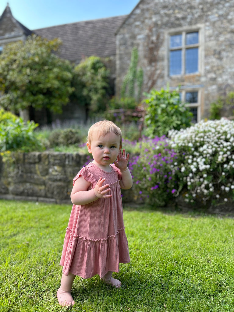 happy toddler stood in front of old stone manor house wearing pale pink two tiered frill dress with frill sleeves