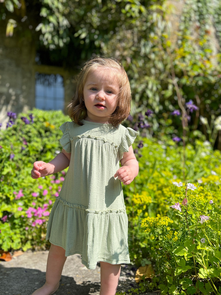 happy toddler standing in front of old stone manor house wearing pale green two tiered frill dress with frill sleeves