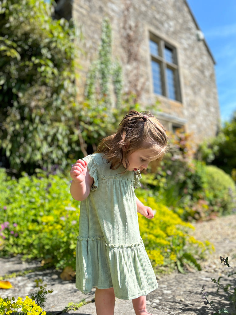 happy toddler standing in front of old stone manor house wearing pale green two tiered frill dress with frill sleeves