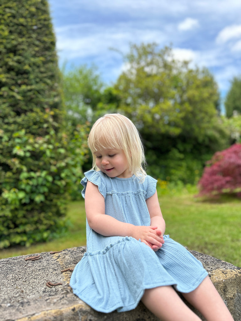 happy girl sat on old stone wall in garden wearing pale blue two tiered frill dress with frill sleeves