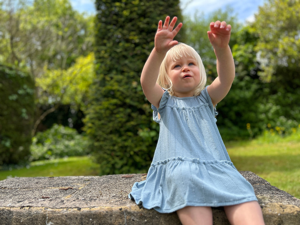 happy girl sat on old stone wall in garden with hands in the air wearing pale blue two tiered frill dress with frill sleeves