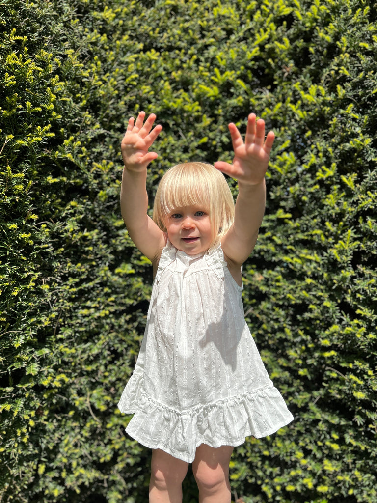 happy toddler standing in sunshine wearing white dress in pretty cutout lace like fabric. Two layer frill sleeves and tier to hem line.