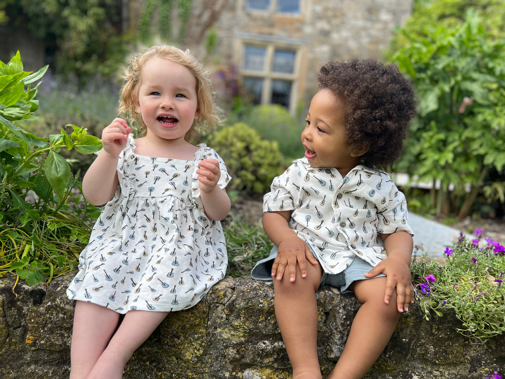 toddler in sunny garden sitting on old stone wall amongst summer plants wearing white dress in cotton jersey with frill over shoulders in guitar print with boy in matching shirt sat next to her