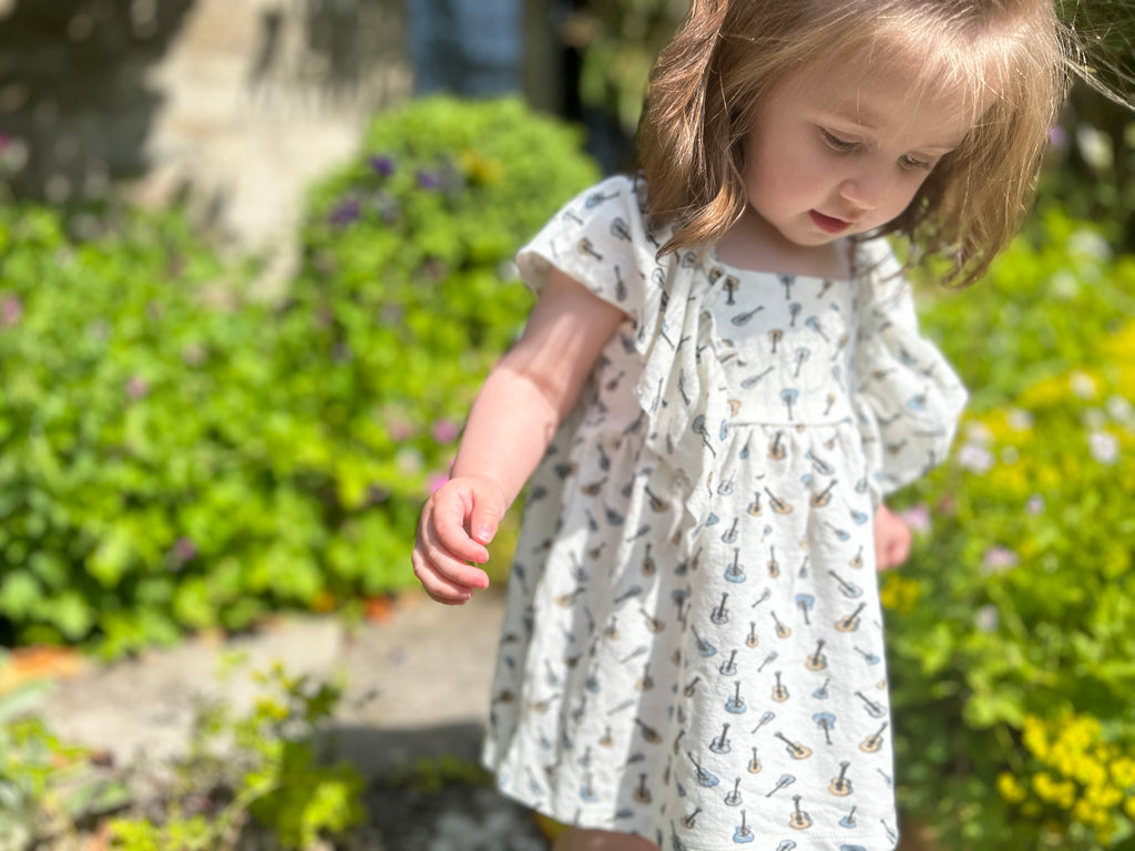 toddler in sunny garden standing amongst summer  plants wearing  white dress in cotton jersey with frill over shoulders in guitar print