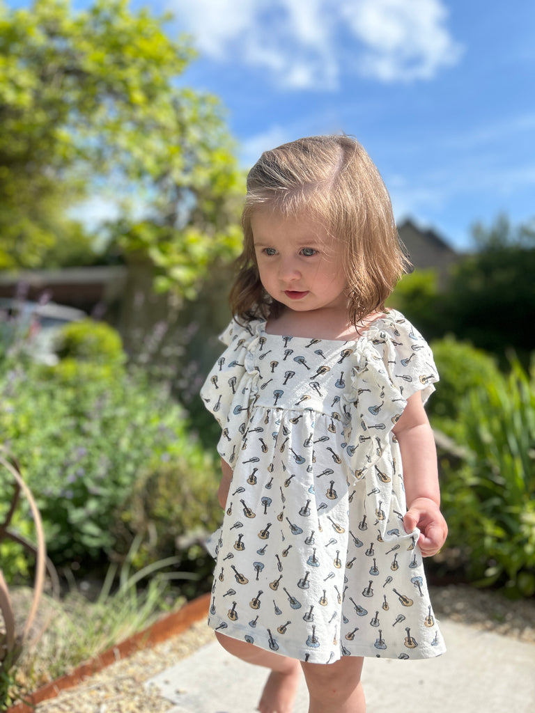 toddler in sunny garden standing amongst summer plants wearing white dress in cotton jersey with frill over shoulders in guitar print