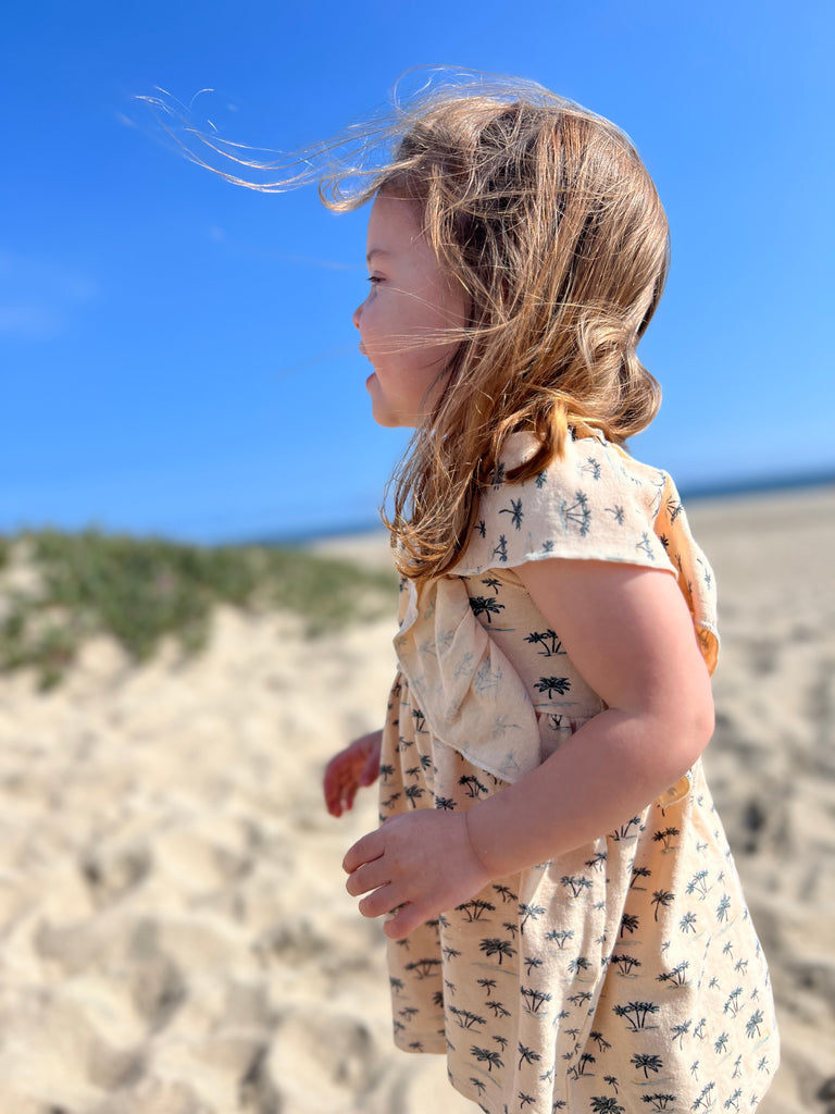 happy girl on sunny beach wearing cream dress in cotton jersey with frill over shoulders and palm tree print