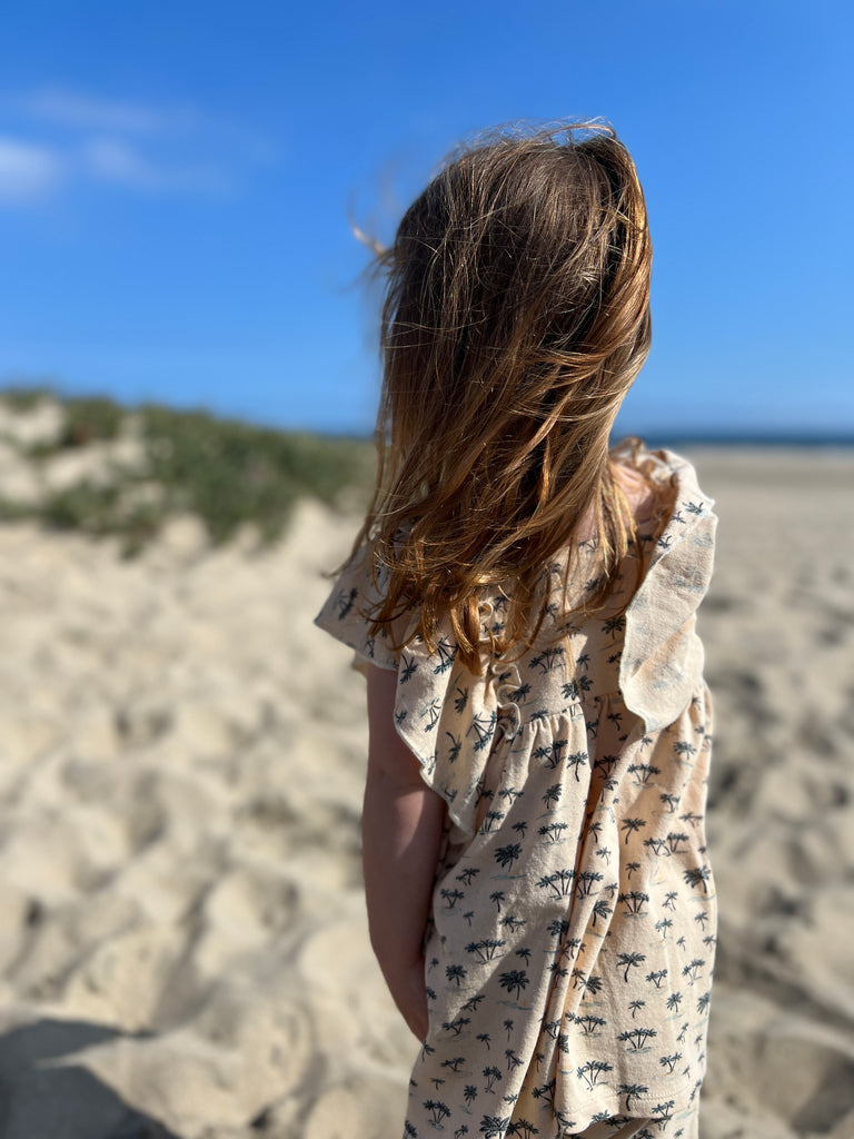 back view of girl on sunny beach wearing cream dress in cotton jersey with frill over shoulders and palm tree print