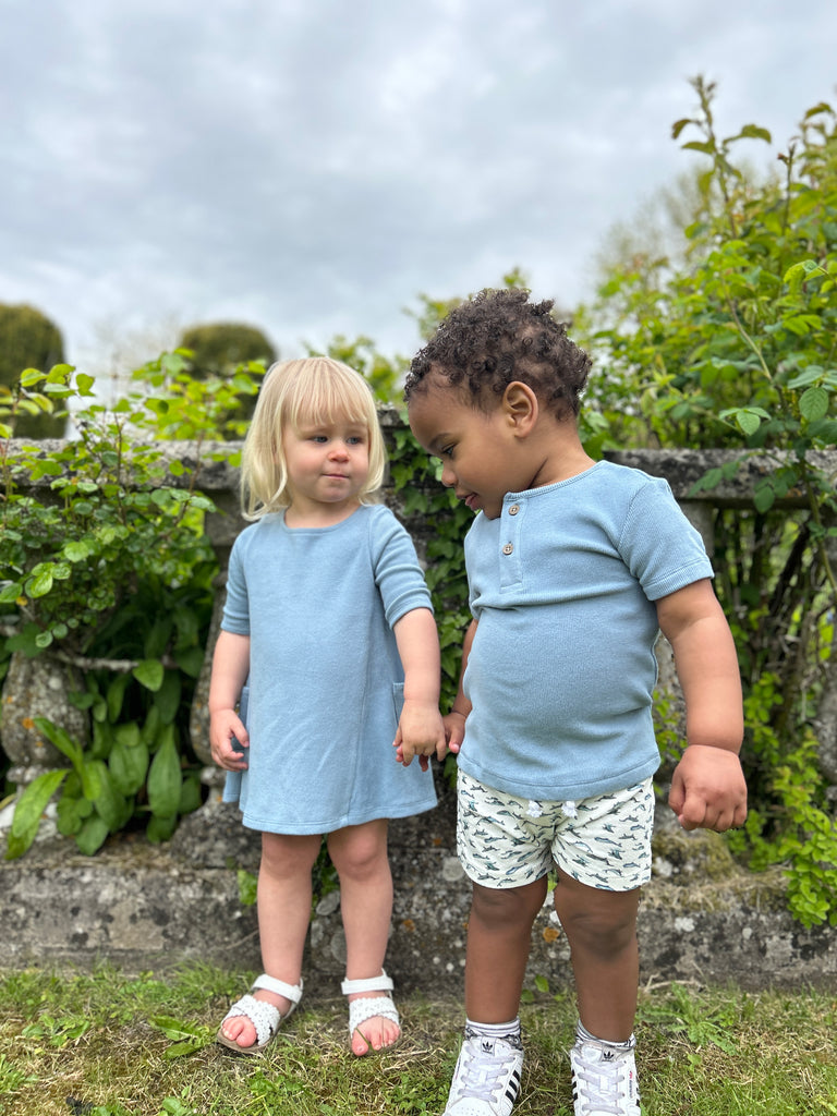 happy toddler in sunny garden wearing pale blue short sleeved henley tee with 2 button detail and whale print shorts girl with matching dress standing at the side