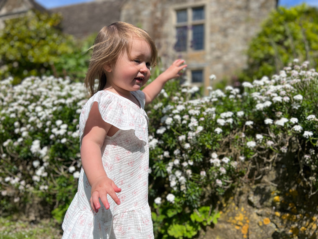 happy toddler in front of white flowers in sunny garden wearing white gauze summer dress with pink flower print. Large over the shoulder frills and v neck front and back