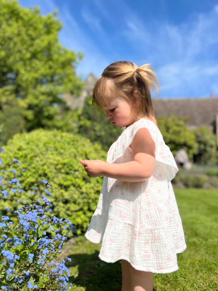 happy toddler in front of blue flowers in sunny garden wearing white gauze summer dress with pink flower print. Large over the shoulder frills and v neck front and back