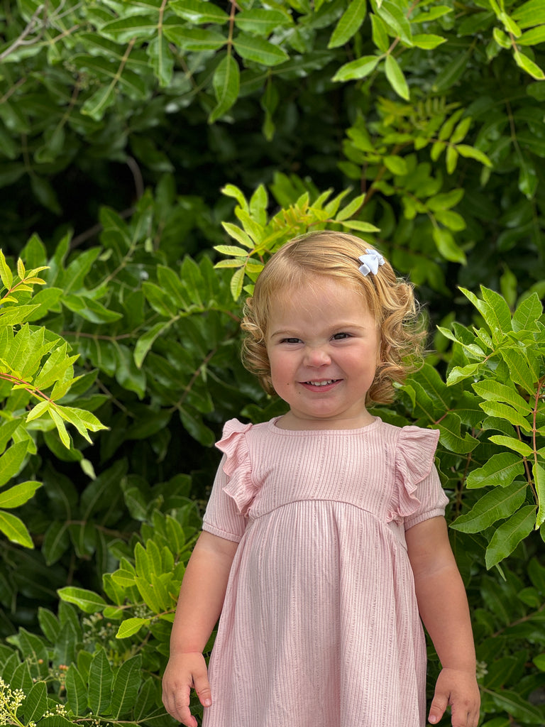 happy toddler standing in front of green bushes on sunny day wearing pale pink dress with frill over shoulders and short sleeves line pattern fabric