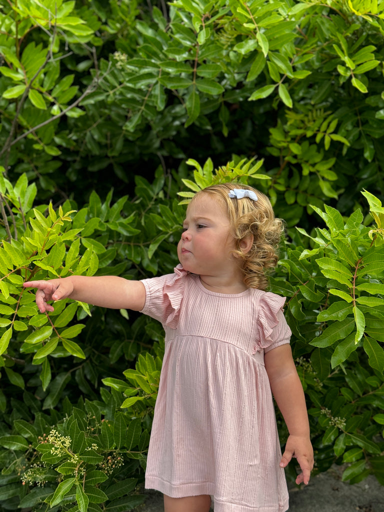 happy toddler standing in front of green bushes on sunny day wearing pale pink dress with frill over shoulders and short sleeves line pattern fabric