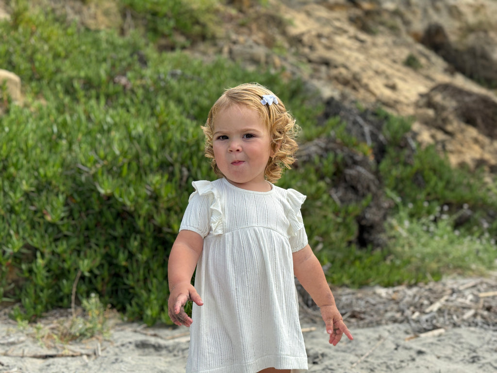 happy toddler standing on beach on sunny day wearing white dress with frill over shoulders and short sleeves line pattern fabric